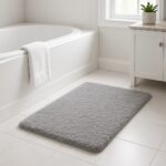 Soft light gray bathroom rug on white tiles next to a bathtub and vanity with a potted plant.